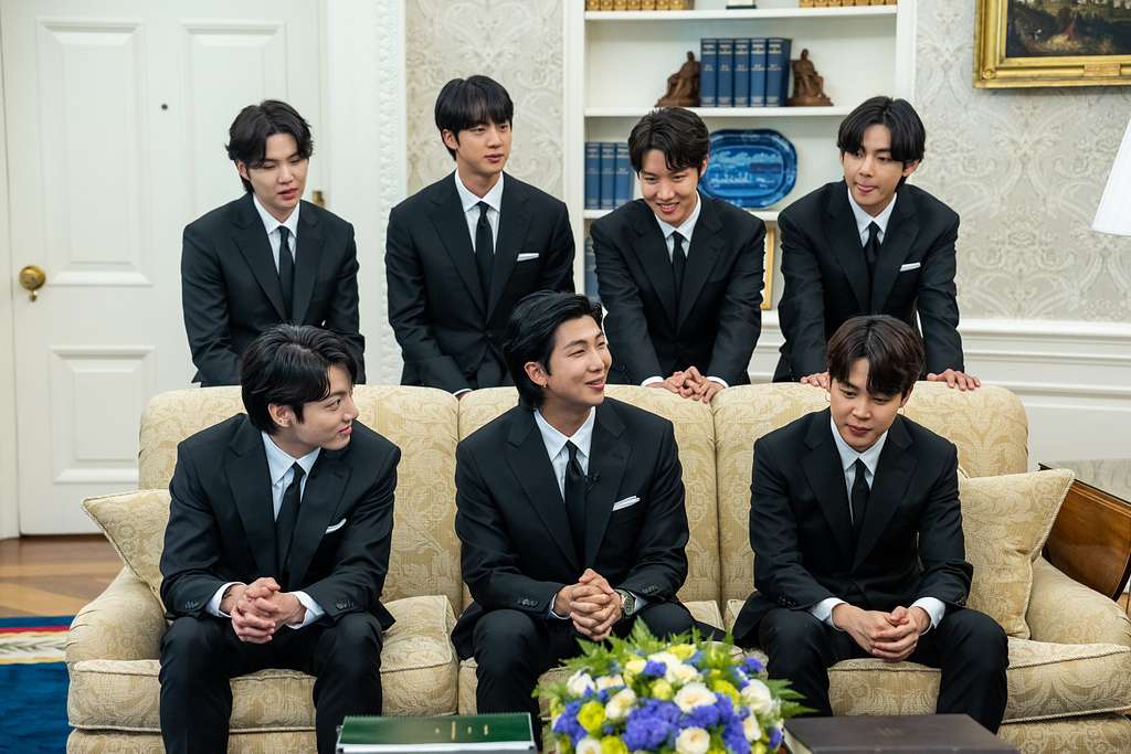 BTS members seated and standing in formal black suits during a public appearance