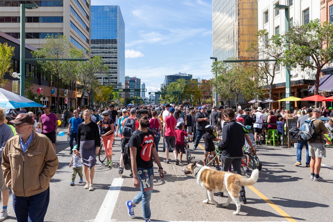 Crowds of people walking along a busy downtown Edmonton street on a sunny day, with office buildings, outdoor patios, and cyclists in the background