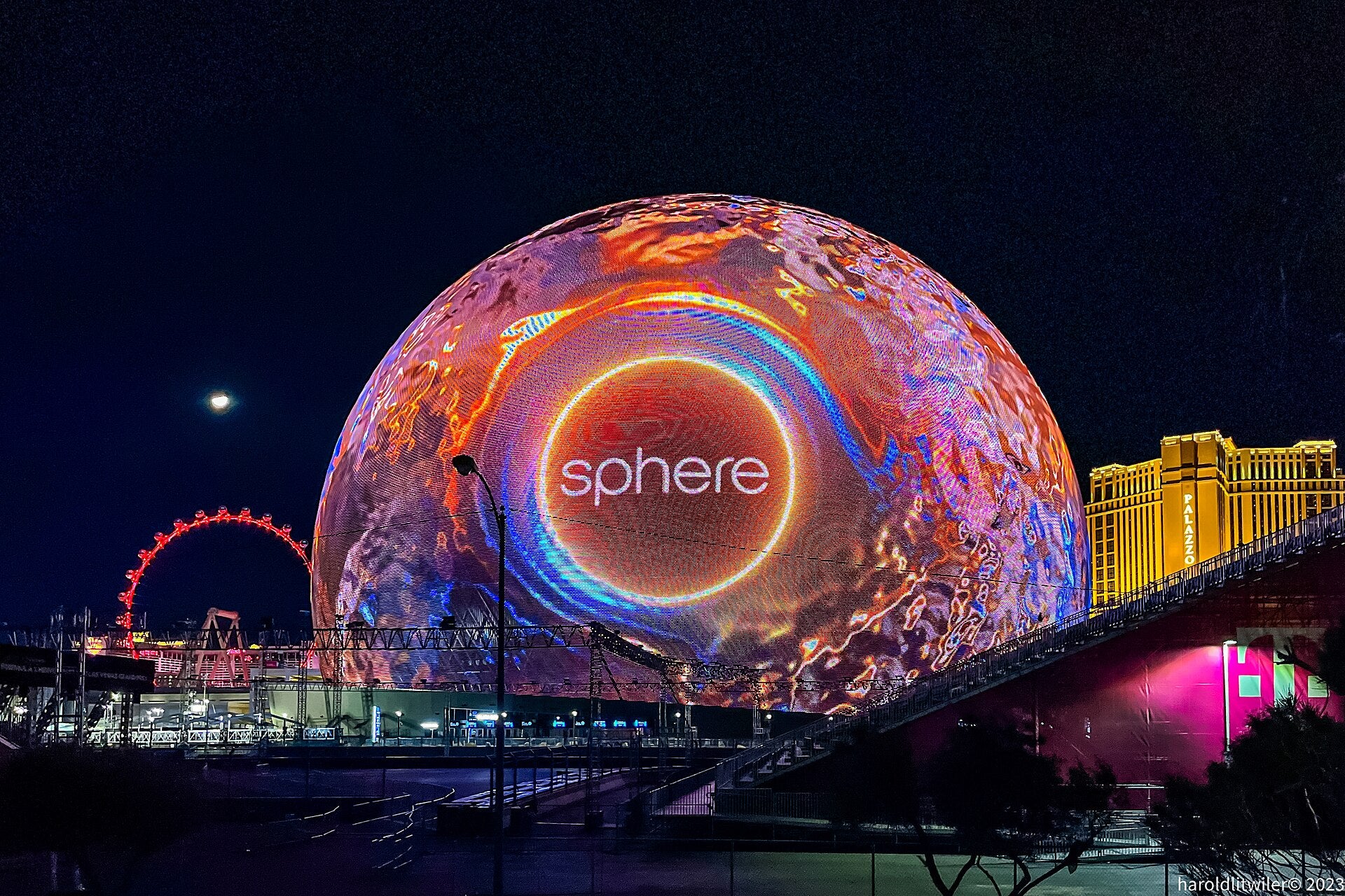 Sphere Las Vegas Adds Rush, Pink, and Metallica Shows The Sphere in Las Vegas lit up at night with a glowing orange and blue display, alongside the High Roller wheel and Palazzo hotel
