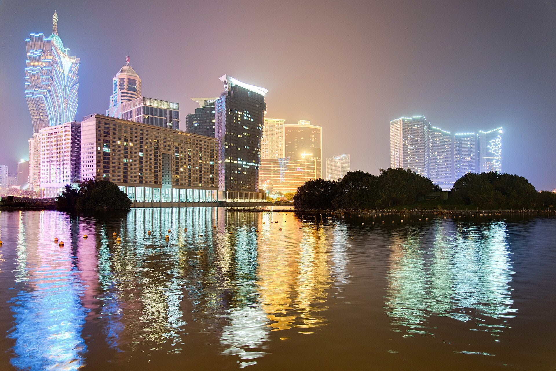 Macau VIP Gaming to Stay Small Share of GGR, IMF Says Macau casino skyline at night, with Grand Lisboa and Wynn Macau lit up and reflected in Nam Van Lake