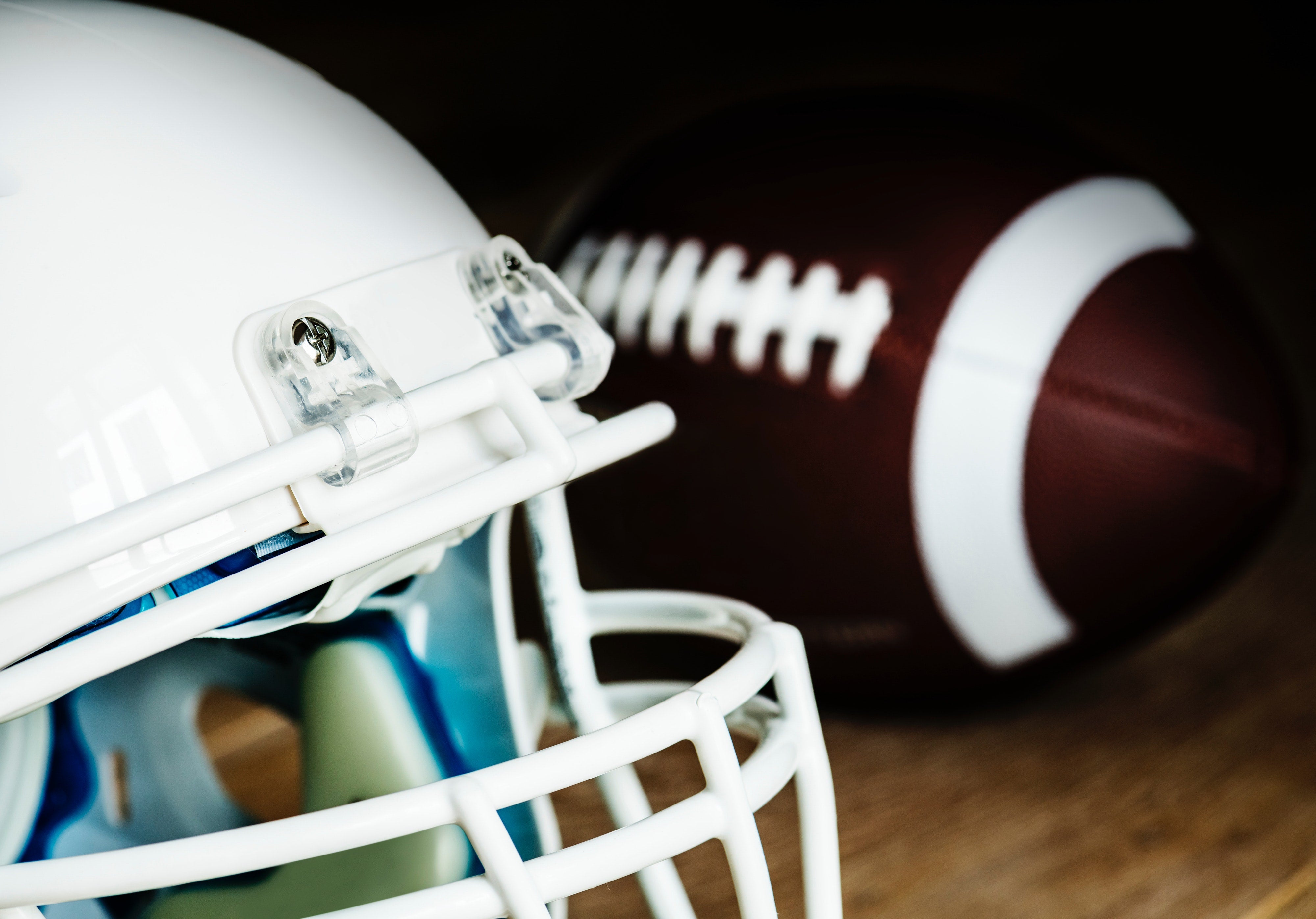 White football helmet with faceguard next to brown leather football on wooden surface