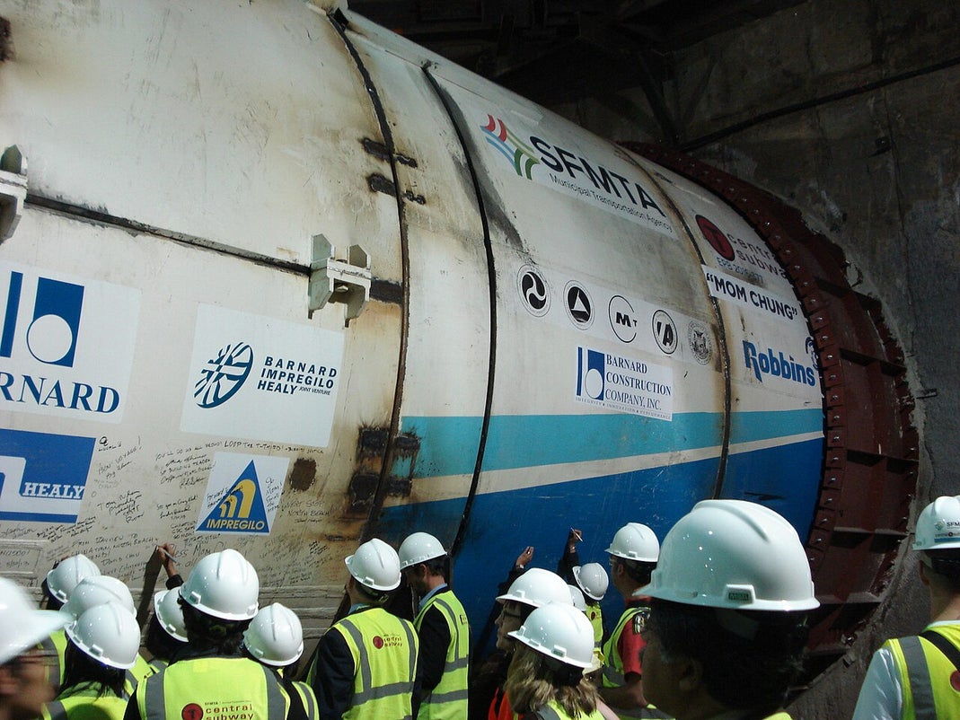 Workers in safety helmets stand beside a large tunnel boring machine during an underground transit tunnel construction project