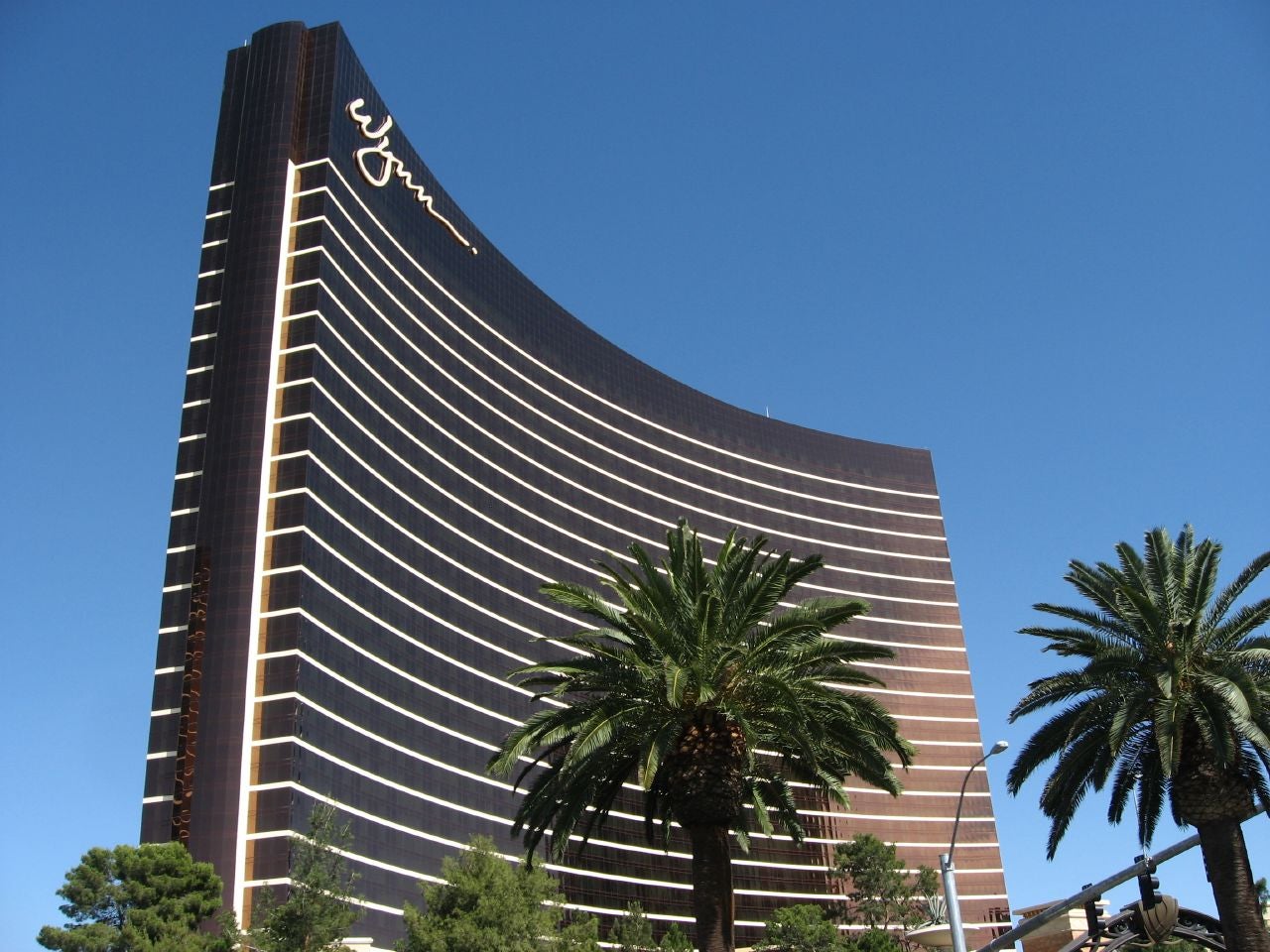 Wynn Resorts Closes $900M Senior Notes Offering Due 2035 Wynn Las Vegas curved bronze glass hotel tower with its signature script logo, framed by palm trees against a clear blue sky