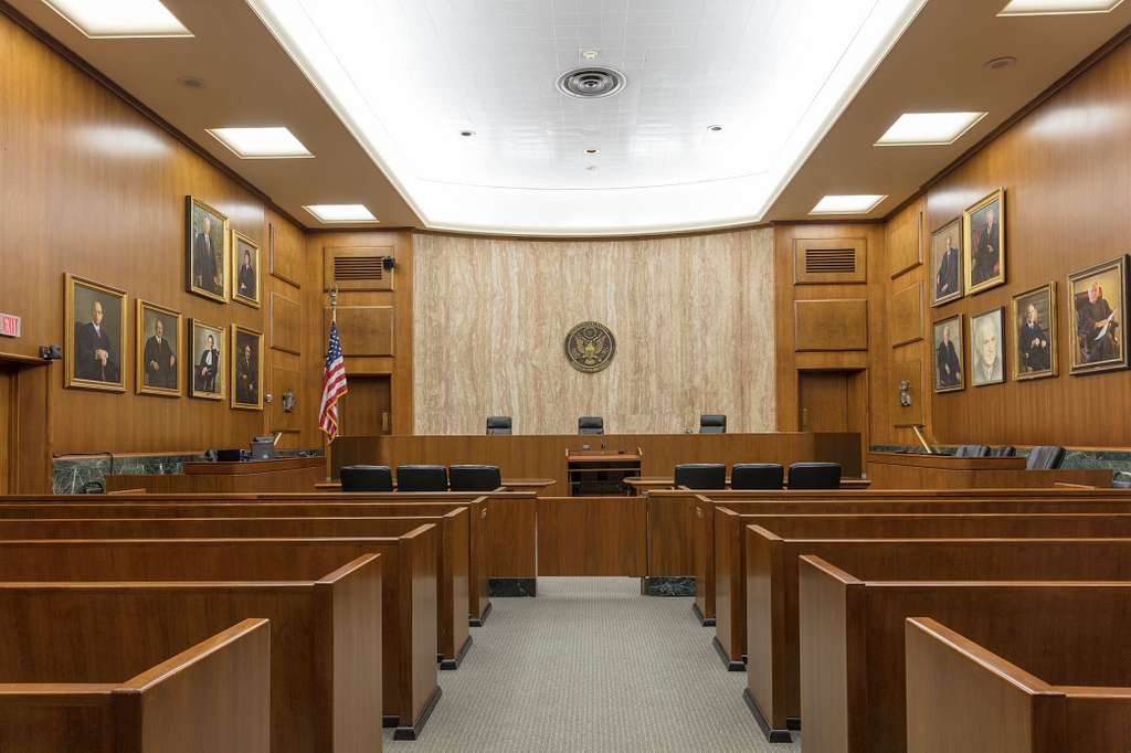 Interior of the E. Barrett Prettyman Federal Courthouse courtroom in Washington, D.C., showing wooden benches, judge’s bench with U.S. seal above, American flag, and portraits of legal figures on the walls
