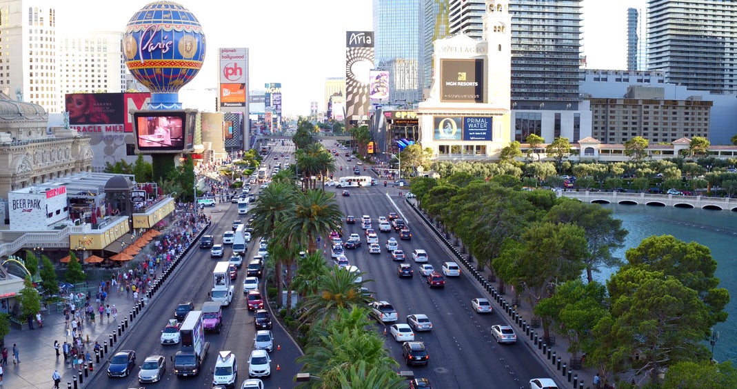 Daytime view of the Las Vegas Strip showing traffic, crowds, and iconic resorts including Paris Las Vegas and the Bellagio