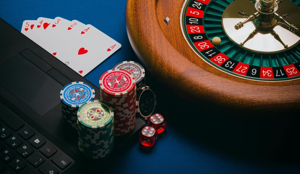 An image of a laptop corner with cards, casino chips and a roulette wheel