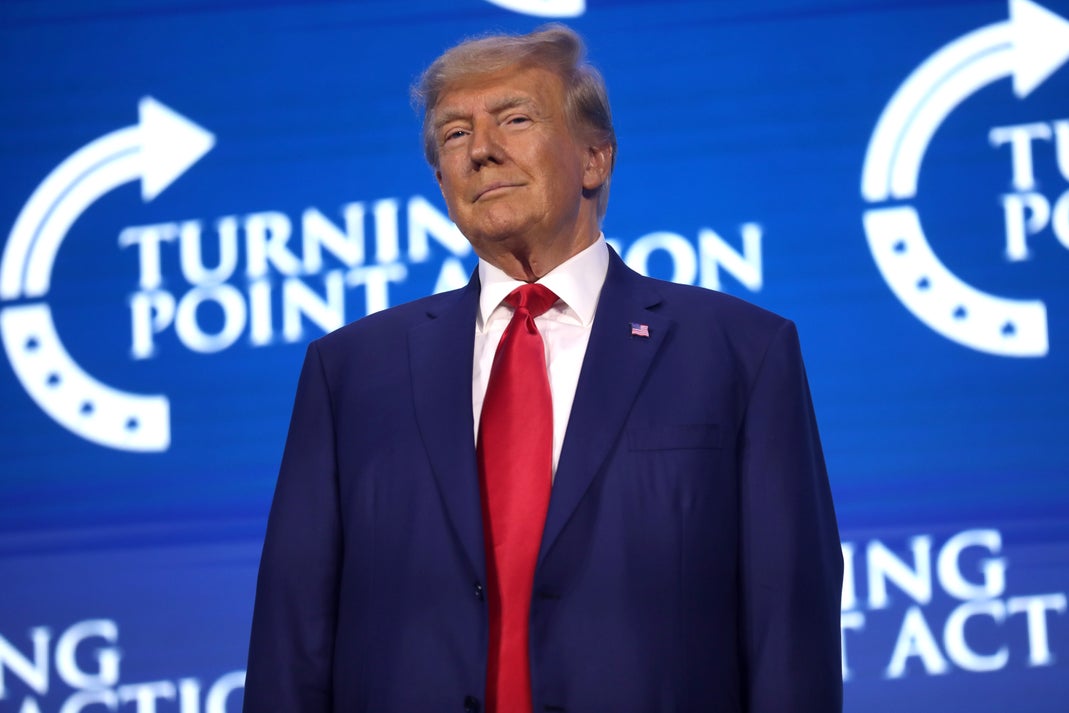 Donald Trump standing at a Turning Point Action event in a navy suit and red tie
