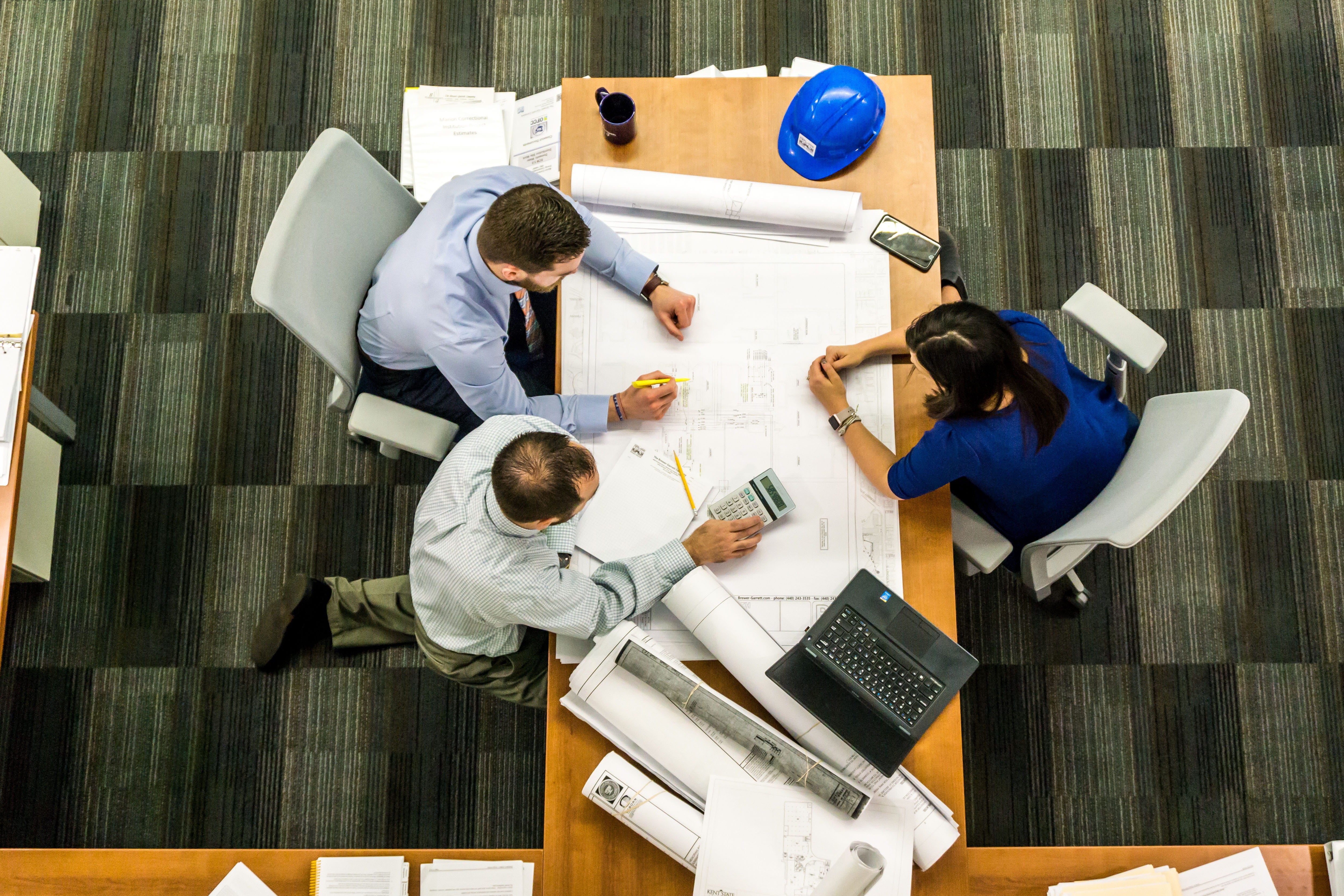 Red Rock Casino Must Bargain With Culinary Union Now Overhead view of three professionals collaborating around a wooden table with blueprints, laptop, calculator, and hard hat during a business meeting