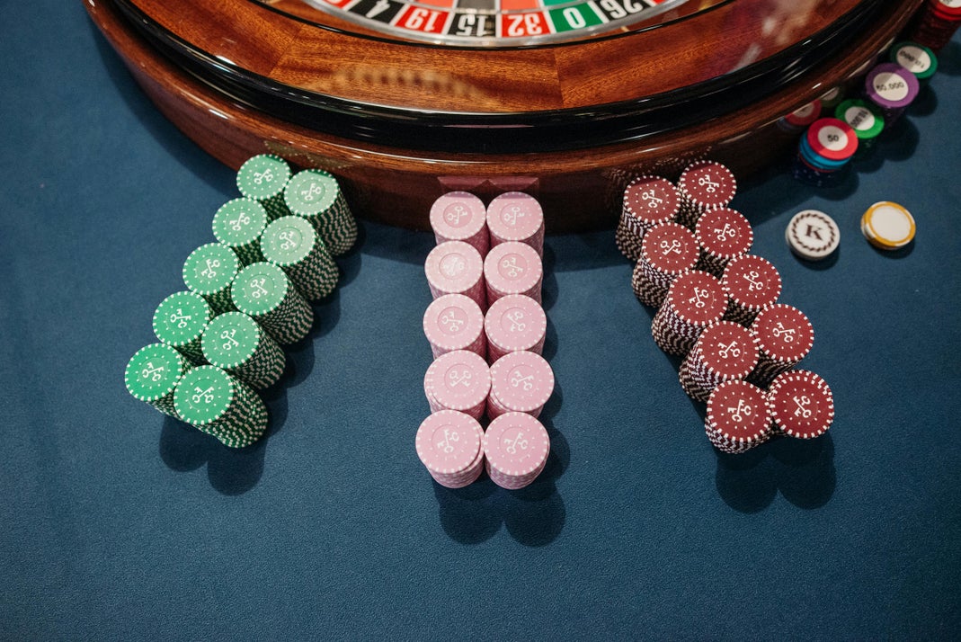 Stacks of green, pink, and red casino chips arranged on a blue felt surface in front of a roulette wheel