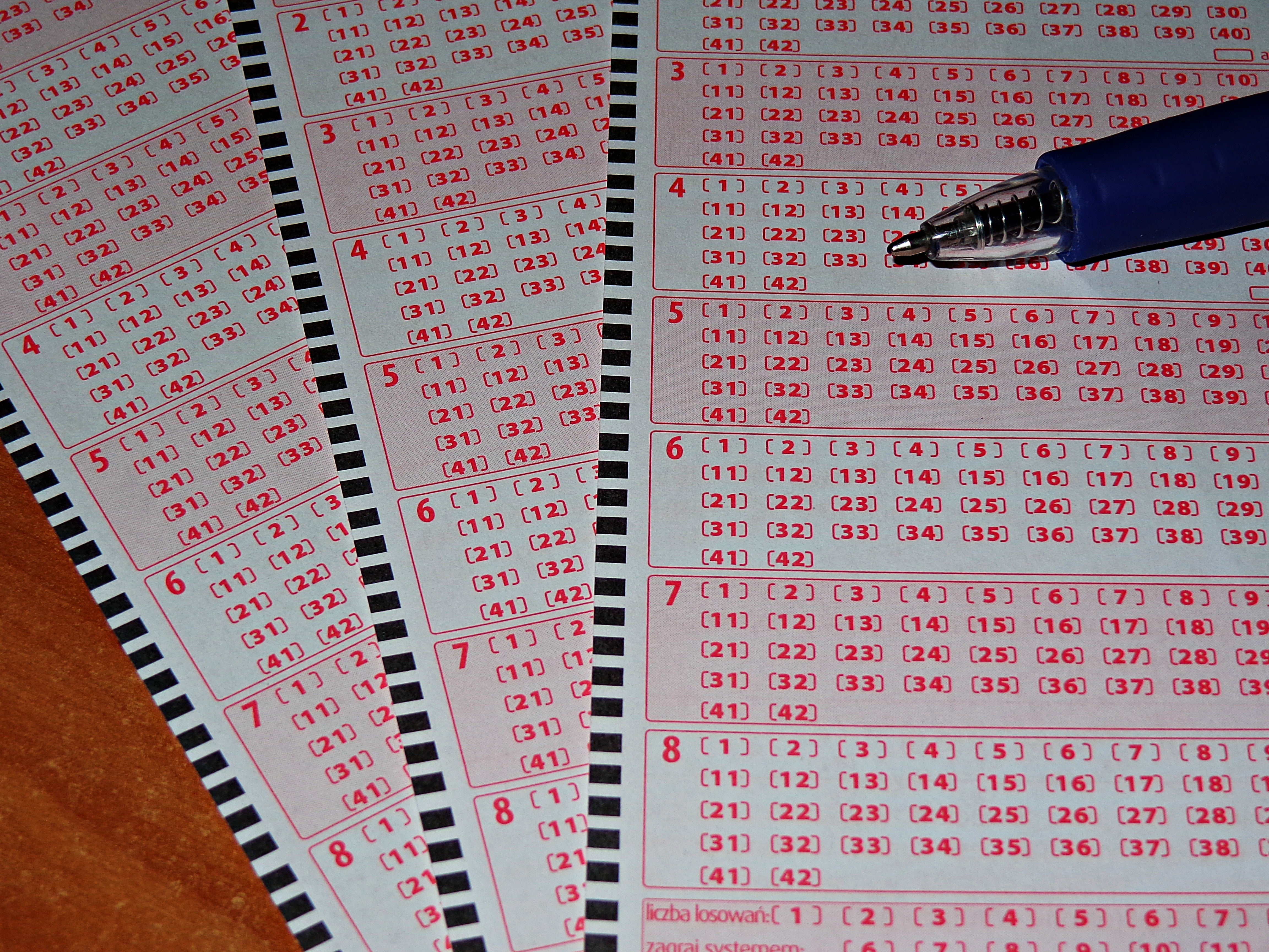 Lottery ticket forms laid out on a wooden surface with a blue pen placed on top, showing numbered grids and barcode strips for lottery selection.
