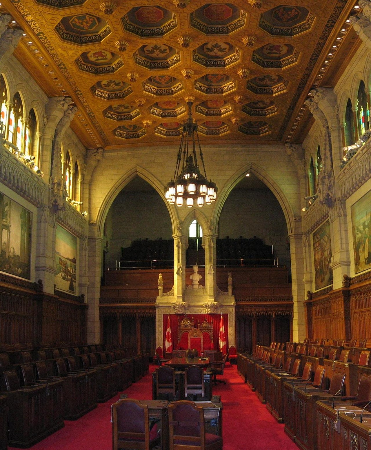Interior of the Senate of Canada chamber showing red carpet, wooden seats, and ornate gilded ceiling in Ottawa