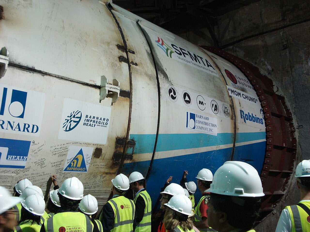 Workers in safety helmets stand beside a large tunnel boring machine during an underground transit tunnel construction project