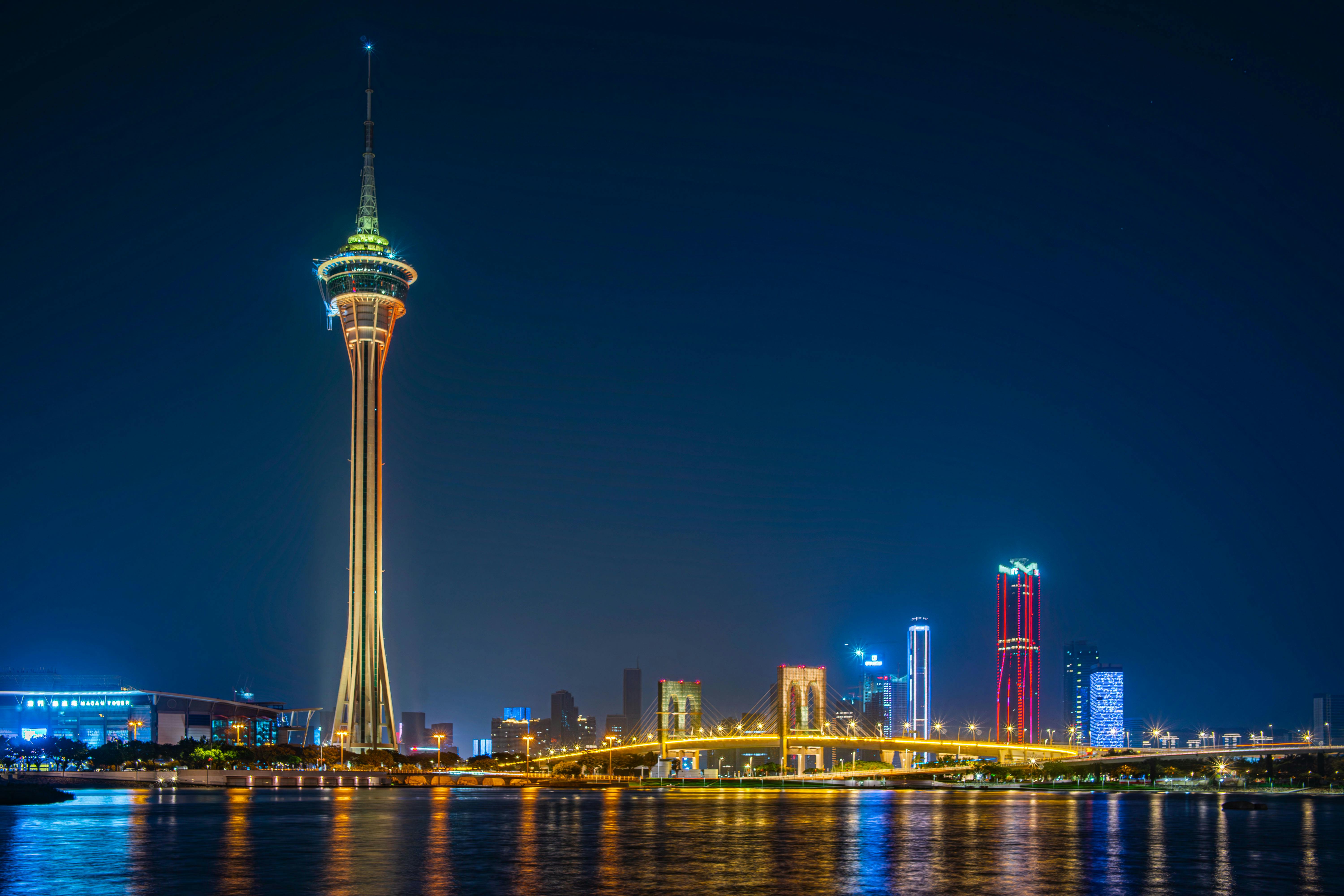 Nighttime view of Macau Tower with illuminated skyline, bridge lights, and reflections on the water