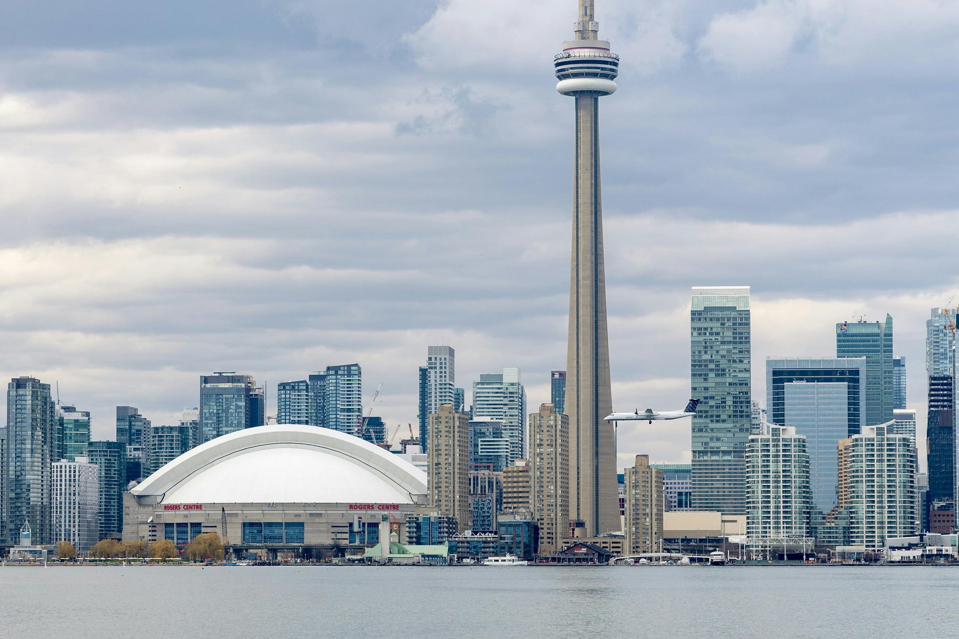 Toronto skyline viewed from the water, featuring the CN Tower and Rogers Centre with a plane flying past