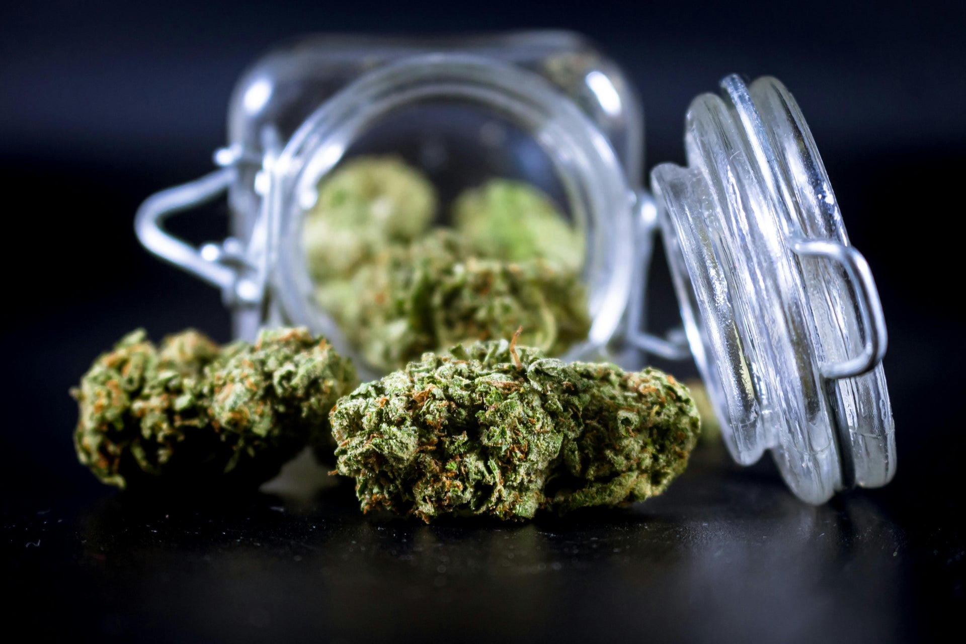 Cannabis buds spilling out of an open glass jar on a dark surface