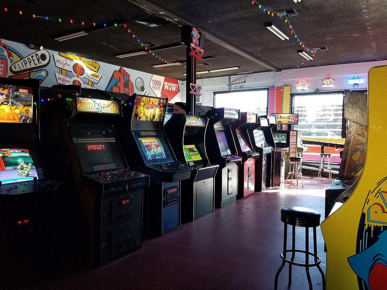 Retro-style arcade with classic game machines, comic wall art, and colorful string lights
