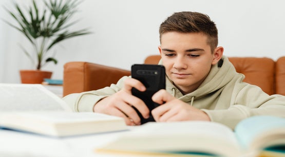 Young male using smartphone with open books nearby Young male looking at smartphone surrounded by open books