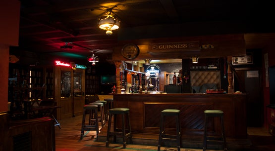 Dimly lit tavern interior with a wooden bar, green bar stools, and neon Budweiser and Guinness signs